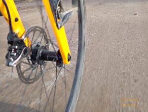A close up of a front wheel on a yellow bike on a chip seal road with the Vulpine tire.