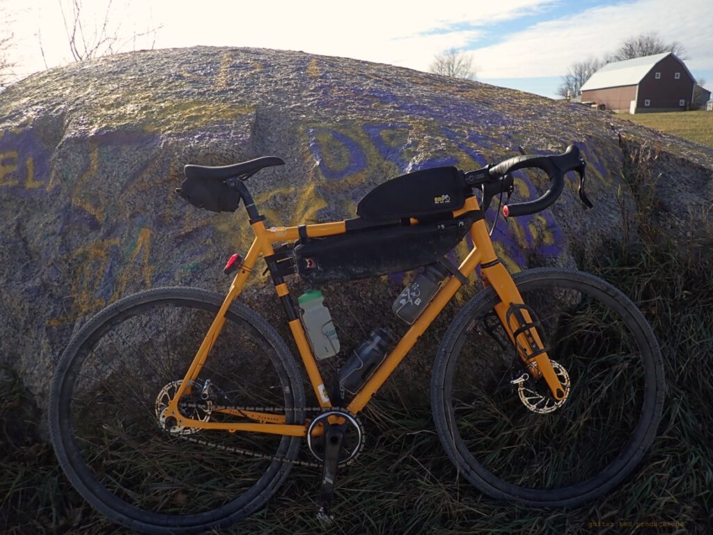 A yellow bicycle with bags leaning against a large boulder in a rural setting